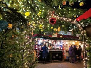 Scorcio notturno di un chiosco in legno da mercatino di Natale riccamente addobbato con ghirlande verdi, palline colorate e archi di micro luci a LED con visitatori davanti al bancone.