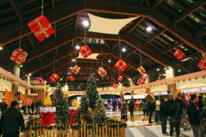 Grande galleria commerciale coperta allestita a festa per il periodo natalizio con grandi pacchi regalo luminosi sospesi al soffitto, alberi di Natale decorati e staccionate in legno.