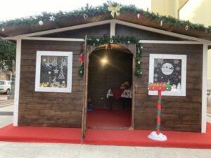Grande casetta di Babbo Natale in legno a noleggio con decorazioni natalizie, festoni e tappeto rosso per eventi e mercatini in piazza.