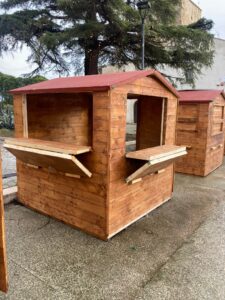 Casetta in legno modello 2x2 metri a pianta quadrata allestita all'aperto in un'area urbana con bancone frontale e laterale a ribalta aperti.