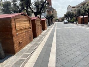 Fila di casette in legno modello 3x2 metri con tetto a due spioventi installate consecutivamente lungo un ampio marciapiede cittadino di giorno.