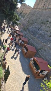 Fila di casette in legno modello 3x2 metri allestite all'aperto lungo un sentiero pedonale ai piedi delle grandi mura di un castello medievale di Termoli.