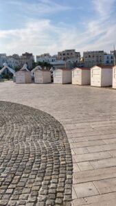 Fila di casette in legno da mercatino modello 3x2 metri installate in semicerchio su una piazza pavimentata con edifici storici sullo sfondo sotto un cielo sereno.