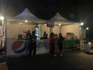 Due gazebo a pagoda bianchi da 4x4 metri uniti e illuminati di notte, allestiti come punto cassa e distribuzione bevande durante un evento all'aperto.