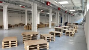 Interno di un ampio spazio industriale della Zecca dello Stato con pile di bancali in legno e pallet standard disposti per un allestimento logistico.