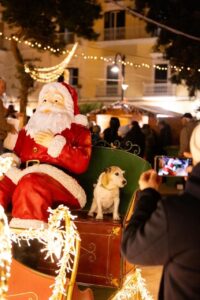 Figura di Babbo Natale seduta su una grande slitta rossa decorata insieme a un cagnolino mentre una persona scatta una foto durante un evento serale.
