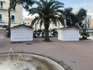 Casette e chioschi in legno bianco per vendite al dettaglio in formato 3x2, posizionati in un'area urbana pavimentata con una fontana, alberi e una palma sullo sfondo