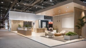 Stand fieristico moderno di grandi dimensioni con pareti bianche, dettagli in legno chiaro e area salotto con poltrone