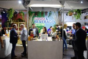 Persone e visitatori riuniti intorno a uno stand fieristico con la scritta luminosa Food and Beverage Expo e decorazioni di piante.